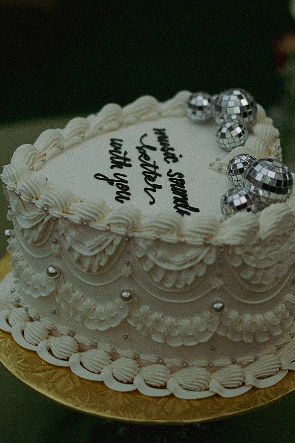 Wedding cake with white buttercream frosting, piped swags and pearl dragees, topped with silver disco balls on a gold stand against dark blur backdrop