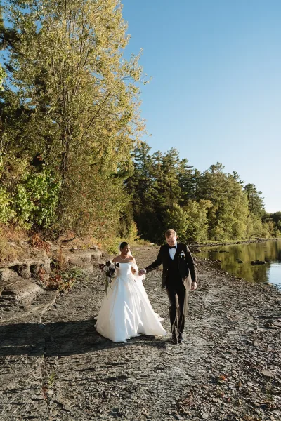 Couple portrait of bride in off-the-shoulder gown and groom in tuxedo holding hands on a rocky lakefront with forest behind
