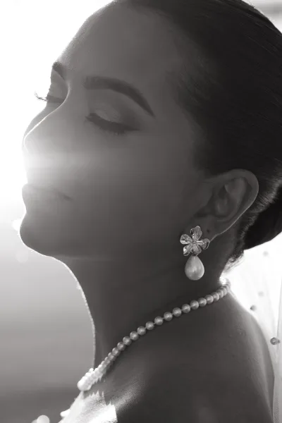 Bridal portrait of a bride in side profile wearing a veil, pearl necklace and drop earrings, with soft glam makeup and sun flare sky backdrop