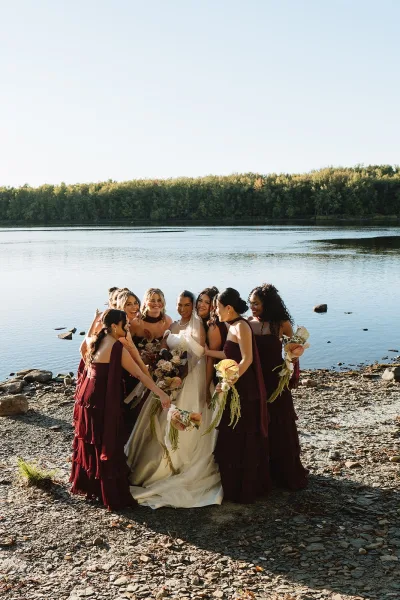 Bride with bridesmaids posing in a close group photo, holding bouquets with veil flowing, on a rocky lakeshore with trees and sky