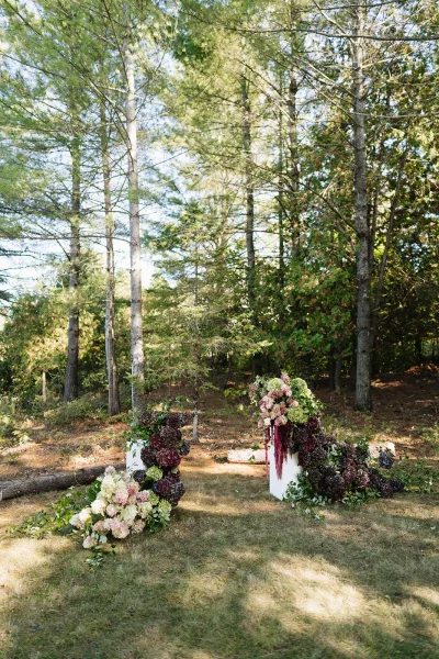 Wedding ceremony backdrop with hydrangea and greenery garland on white pedestals, burgundy hanging blooms in a forest clearing
