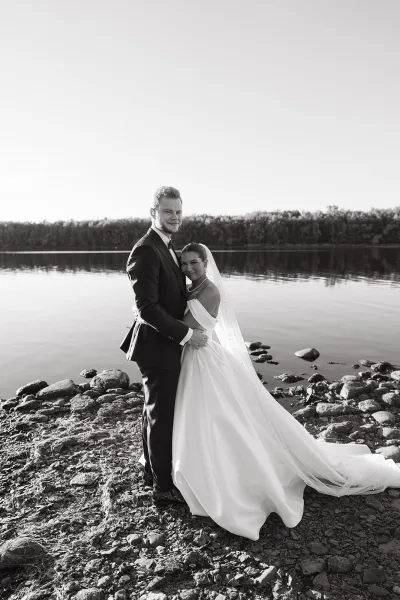 Couple portrait in a black and white wedding portrait style, bride in veil and gown embracing groom in tuxedo by a lakeside rocky shore