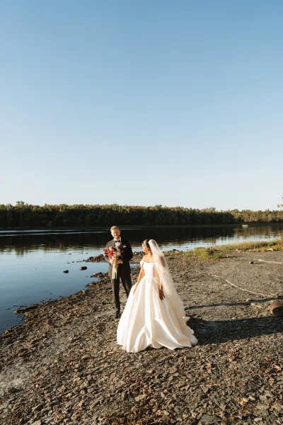 Couple portrait of bride in strapless ball gown and long veil with groom in black tuxedo holding bouquet on rocky lakeshore under dramatic sky
