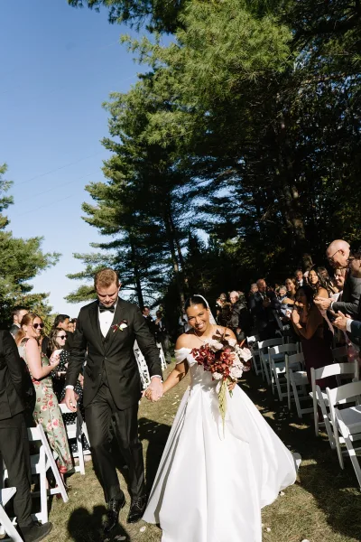 Wedding recessional as bride and groom walk the aisle holding hands, bride with burgundy bouquet and veil on a pine-lined lawn under blue sky