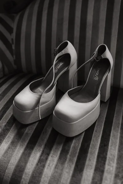 Bridal shoes, white platform block-heel ankle-strap heels resting on a striped upholstered chair during bridal prep details