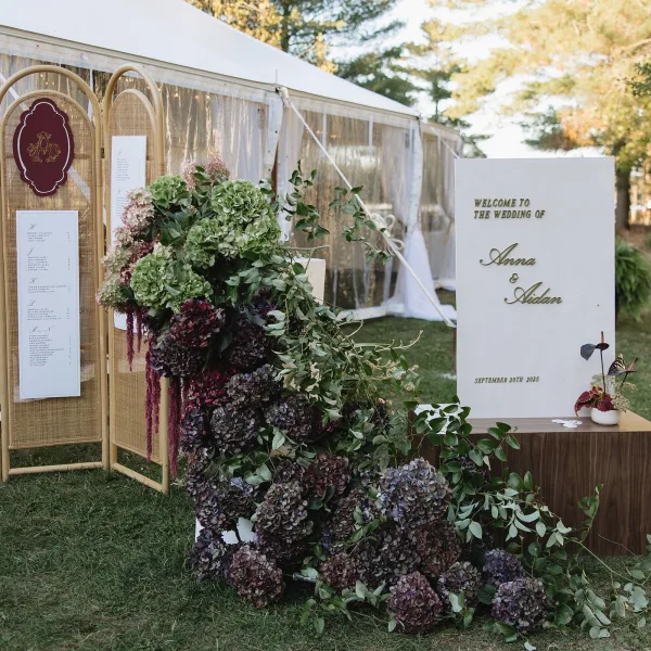 Wedding welcome sign with gold lettering and hydrangea florals beside a rattan seating chart under a clear-top tent on grass lawn