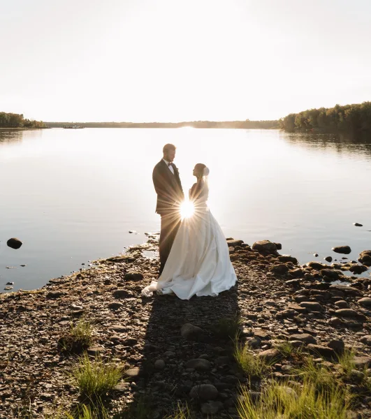 Couple portrait of bride and groom silhouette facing each other on a rocky lakeshore, veil and tuxedo boutonniere glowing in sunset light