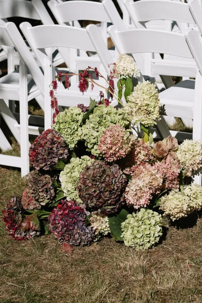 Ceremony aisle florals with hydrangeas, roses, and cascading greenery at the end of a row of white folding chairs on a grass lawn