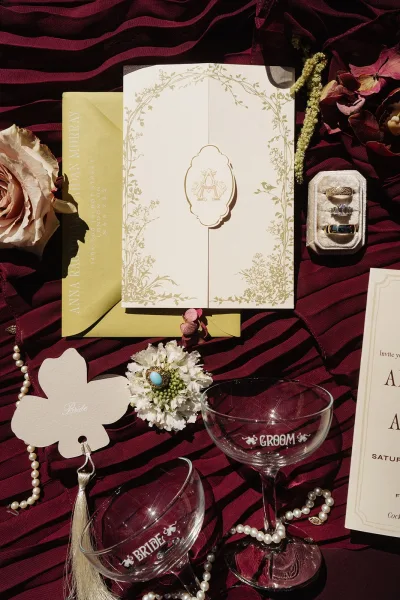 Invitation suite wedding invitation flat lay with wax seal, monogram envelopes, rings, pearls, rose and place cards on burgundy fabric backdrop