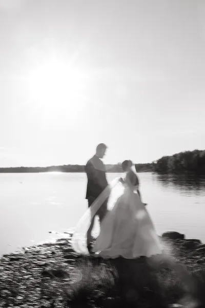 Couple portrait of bride and groom by lake, her veil blowing in the wind as sun flare glows over shoreline rocks and trees