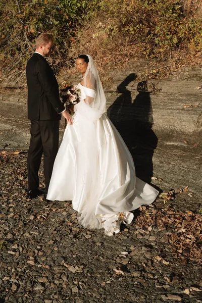 Couple portrait of bride and groom holding hands, her cathedral veil and bouquet flowing beside a boulder amid autumn trees.