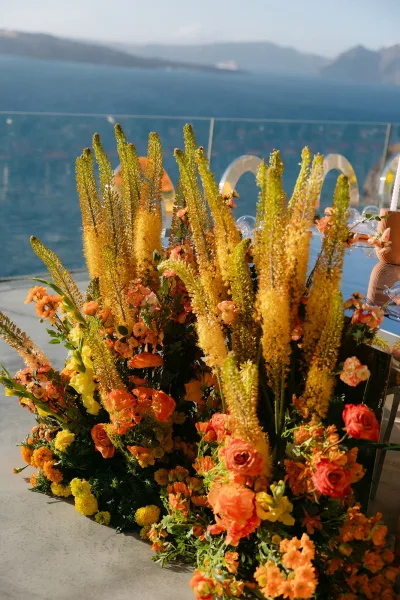 Wedding floral installation featuring orange and yellow wedding flowers with acrylic chairs, on a terrace overlooking the ocean and mountains.