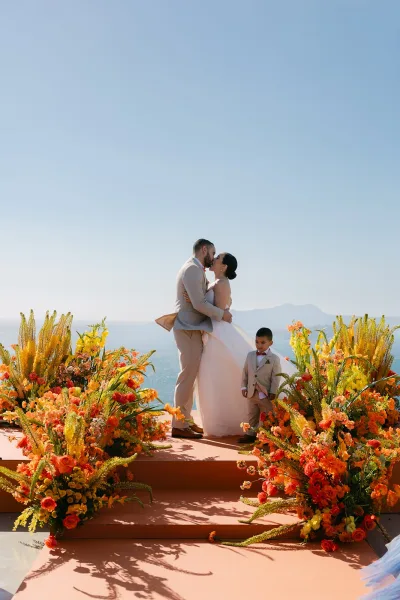 Wedding first kiss as bride and groom embrace amid orange and yellow florals, with ocean, blue sky, and distant mountains behind the altar.