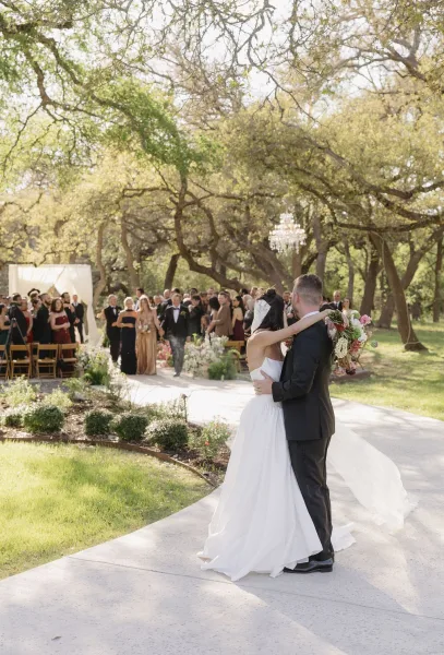 Wedding recessional with newlyweds walking down the aisle, bride's bow veil trailing as they exit along a concrete pathway amid garden trees.