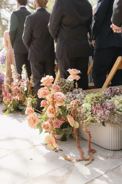 Ceremony aisle flowers shape a garden ceremony floral design, with roses cascading along a sunlit stone patio framed by garden trees.