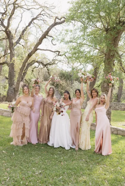 Bride and bridesmaids in mismatched bridesmaid dresses stand together on a grass lawn by a stone wall framed with trees.