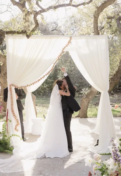 Wedding first kiss captured in an outdoor ceremony moment beneath a draped ceremony arch, on a stone patio framed by trees.