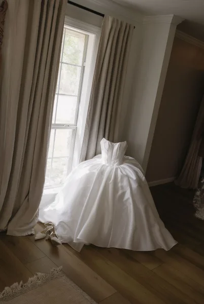 Wedding dress close-up of a strapless wedding gown with a voluminous skirt, bridal shoes peeking beneath it by a sunlit window.