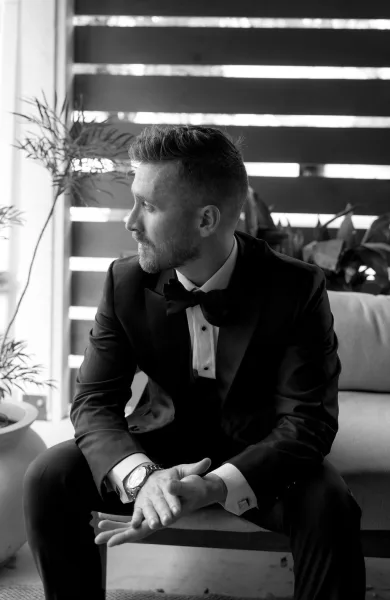 Groom portrait in black and white, seated with a bow tie against a wooden slat wall and potted plants beside a sofa.