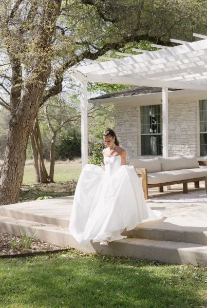 Bridal portrait of a bride holding her dress as she walks down patio steps, drop earrings catching light, beside a stone house and pergola.