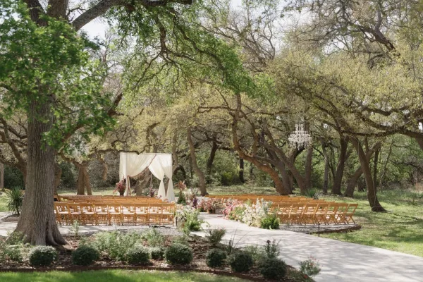 Outdoor ceremony setup featuring garden ceremony decor with a draped arch on a lawn, framed by trees and a winding garden path.