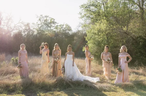 Bridal party portrait of a bride with bridesmaids in mix and match blush and champagne dresses, standing in sunlit meadow grass framed by trees.