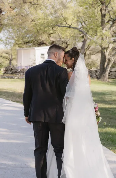 Couple portrait of a bride and groom walking along a tree-lined path by a stone wall, her long veil trailing as they share a quiet, romantic moment.