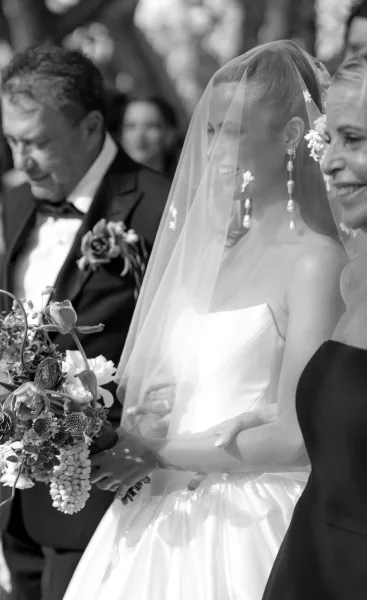 Ceremony processional with a bride walking down aisle beneath a blusher veil, guests flanking sunlit garden trees in the background