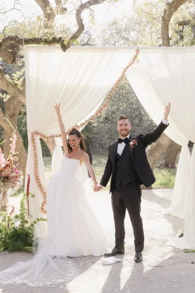 Ceremony moment of a just married celebration as the couple holds hands beneath a draped fabric arch, cheering on a sunlit stone patio.