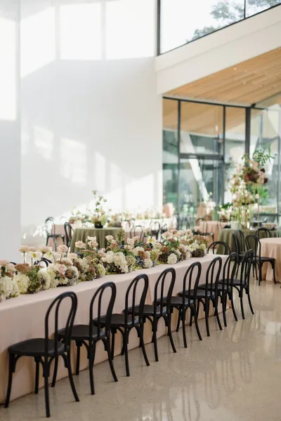 Reception tablescape with a long banquet table wedding setup, blush tablecloth and floral garland centerpiece in a modern hall with big windows