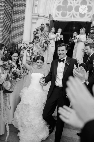 Wedding recessional as newlyweds walk out of a brick church, bride in strapless ruffled gown lifting bouquet overhead amid cheering guests