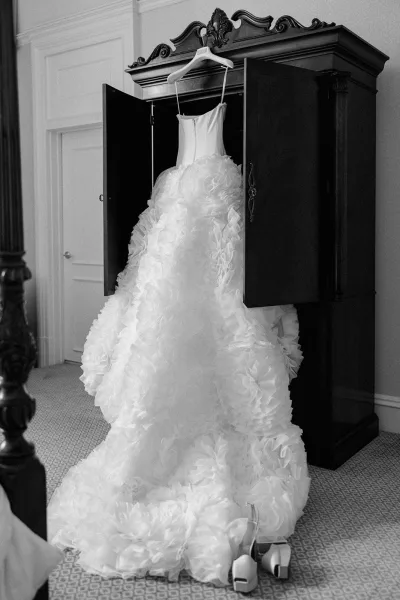 Wedding dress strapless ball gown on a hanger in a bedroom armoire, floral ruffle skirt cascading beside bridal heels on carpet