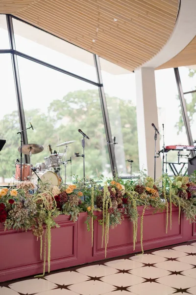 Wedding band stage with microphone stands, drum kit and keyboard, framed by a lush rose and hydrangea floral garland in front of large windows