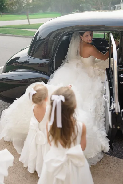 Bride in car in a vintage wedding car, smiling with bouquet as flower girls in white dresses help by a suburban driveway