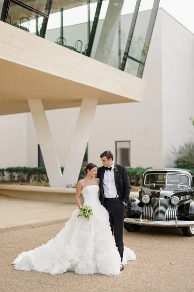 Couple portrait of bride in strapless gown with long train and bouquet beside groom in tuxedo by a vintage car outside glass building entrance