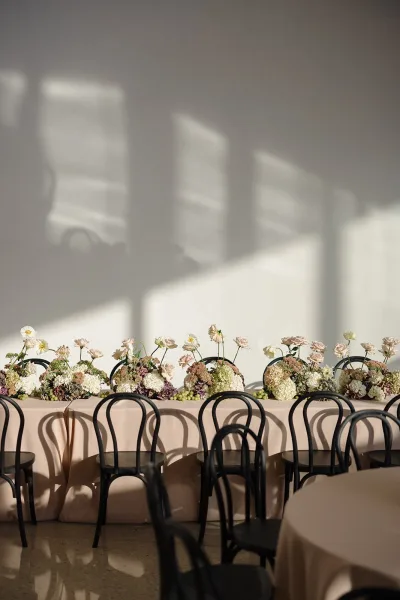 Reception tablescape with head table decor on a long beige banquet table, low hydrangea and blush florals, greenery, and green grapes in soft window shadows