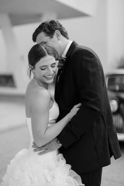Couple portrait in a black and white wedding portrait as groom kisses bride’s forehead, her strapless tulle gown and pearls on a city street
