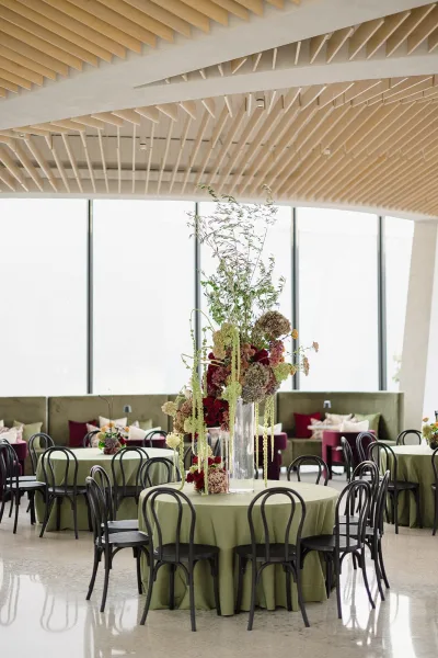 Reception tablescape with tall wedding centerpiece in a glass vase on round tables with green linens, black bentwood chairs, and windowed room