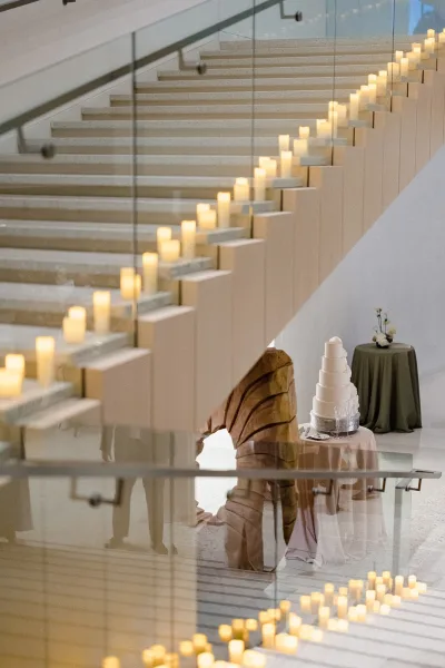Wedding dessert table with tiered wedding cake table decor, candles and champagne flutes on a round linen table by a glass staircase railing