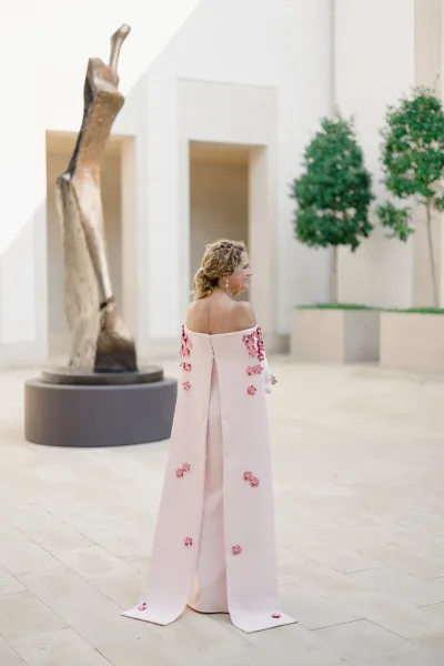 Wedding guest dress in pastel pink with off-the-shoulder cape sleeves and 3D floral appliques, styled with drop earrings in a modern courtyard
