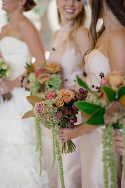 Bridesmaid bouquets with cascading bridesmaid bouquet blooms of roses, hydrangea, and greenery, held by neutral dresses indoors with blurred guests