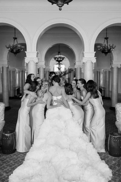 Bride with bridesmaids in a group hug, long train flowing, laughing together in an arched hallway with chandeliers and columns