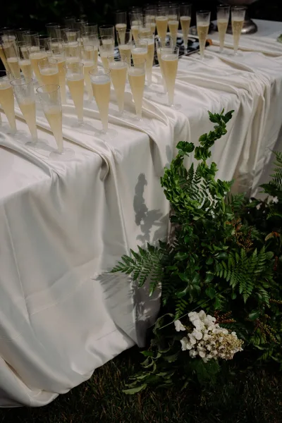 Champagne display at a wedding champagne bar, accented with white florals, set on a grass lawn amid lush garden foliage.