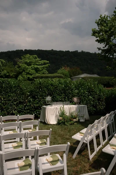 Ceremony seating in a garden ceremony setup with rows of white folding chairs on a hedge-lined lawn, framed by trees and a distant hillside.