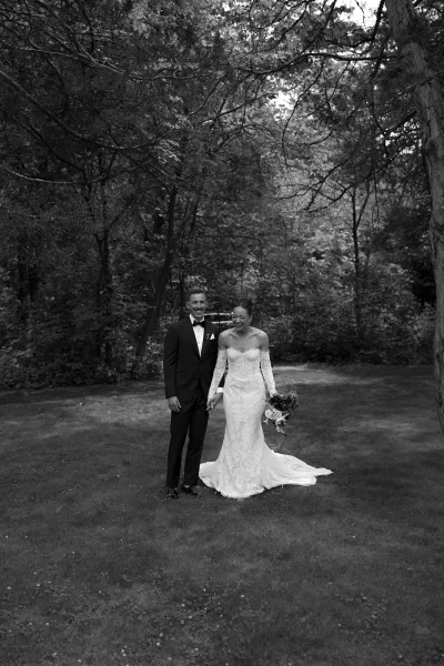 Couple portrait during an outdoor wedding portrait: bride and groom laugh, holding hands, her bridal bouquet bright against woodland trees.