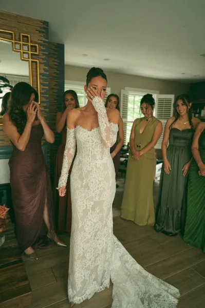Bridesmaid first look as the bride with bridesmaids is revealed in a lace wedding dress, framed by window shutters in an indoor room.