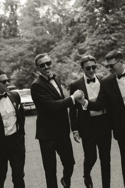 Groom and groomsmen captured in a groomsmen walking photo stride down a tree-lined road in formal attire with sunglasses.