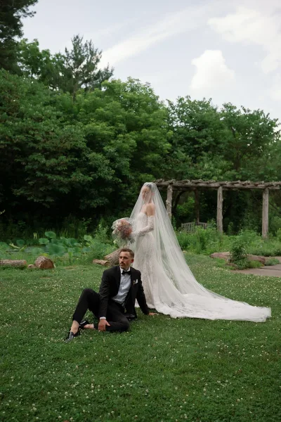 Couple portrait of a bride and groom outdoors under a wooden pergola by a lily pond, her long veil drifting as they pose together.