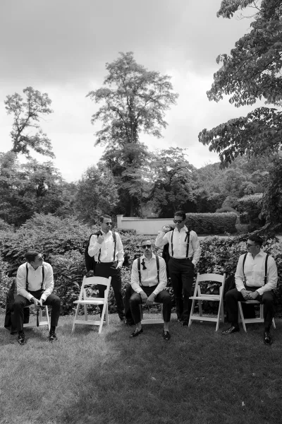 Groomsmen portrait in black-and-white, wearing suspenders, posed on a grass lawn before garden hedges beneath a softly clouded sky.
