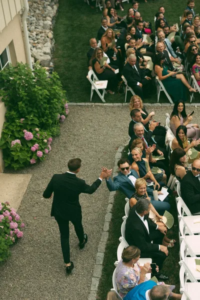 Groom processional as the groom walking down the aisle high-fives cheering guests beside white ceremony chairs, amid garden hydrangeas.
